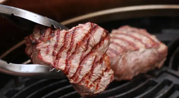 Zangen halten ein saftiges Stück Roastbeef über einem Grillrost. Das Fleisch hat schöne Grillstreifen und ist perfekt gegrillt.