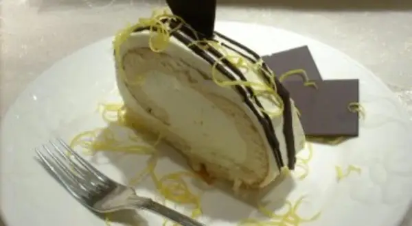 Ein Stück Zitronenrolle mit cremiger Füllung, dekoriert mit Schokoladenglasur und Zitronenzesten. Ein perfektes Zitronenrolle Rezept zum Nachbacken.