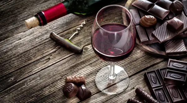 Ein Glas Rotwein und verschiedene Schokoladenstücke auf Holztisch. Ein Korkenzieher liegt daneben.