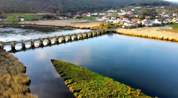 Un puente de piedra con arcos cruza un río azul, conectando un pueblo pintoresco con campos verdes. El puente, conocido como Ponte nafonso, se alza majestuoso sobre el agua.