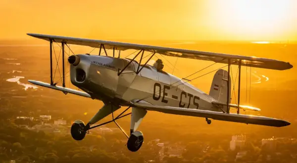Ein silberner Doppeldecker fliegt im goldenen Abendlicht über einer Stadt. Ein Traum für alle, die gerne flugzeuge fotografieren.