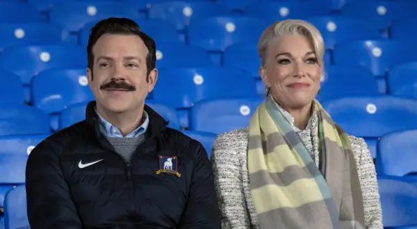 Die Besetzung von Ted Lasso, Jason Sudeikis und Hannah Waddingham, sitzt lächelnd auf blauen Tribünen.