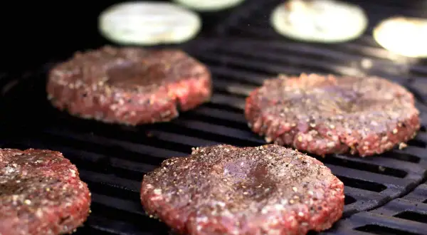 Vier rohe Burger-Patties, die darauf warten, dass man sie selber macht, brutzeln auf einem Grill. Zwiebelringe liegen im Hintergrund.