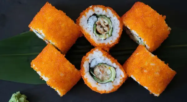 Roladki sushi California z pomarańczowym tobiko, ryżem, awokado i krewetką. Obok zielony listek i odrobina wasabi.