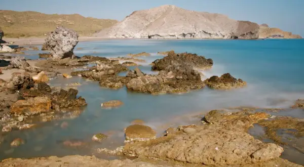 Rocas volcánicas emergen de las aguas tranquilas de la cala de la media luna, con montañas áridas al fondo bajo un cielo azul.