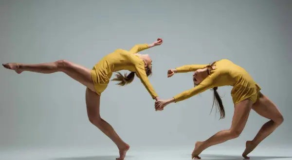 Deux danseuses en tenue jaune, mains jointes, en mouvement synchronisé sur fond gris.