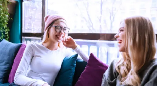 Dos amigas ríen sentadas en un sofá, como si compartieran anécdotas de couchsurfing que es una forma de viajar.