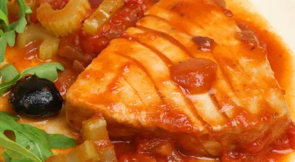 Un succulento tonno in padella con pomodoro, olive nere e sedano, guarnito con rucola fresca.