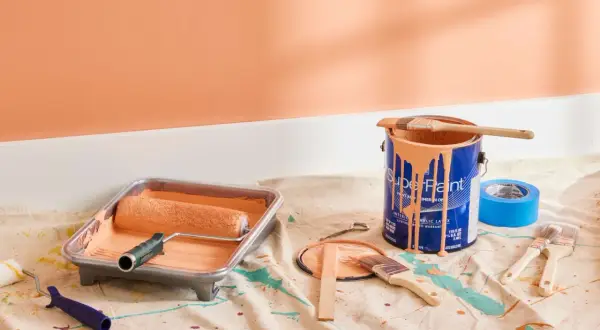 Painting supplies, including a roller in a tray and a can of peach-colored satin paint, are laid out on a drop cloth.