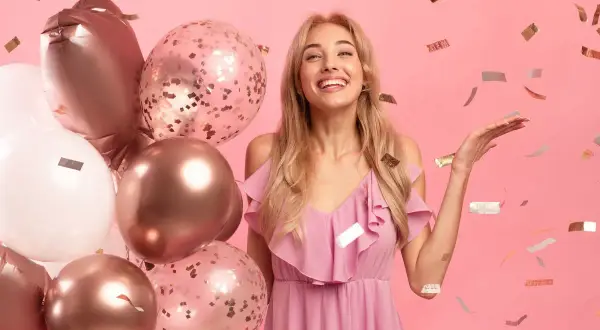 Radosne życzenia urodzinowe dla bratowej! Uśmiechnięta blondynka w różowej sukience, otoczona balonami i konfetti.