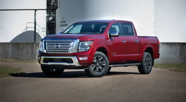 Czerwony Nissan Titan z chromowanymi detalami stoi na asfalcie przed białymi zbiornikami przemysłowymi.