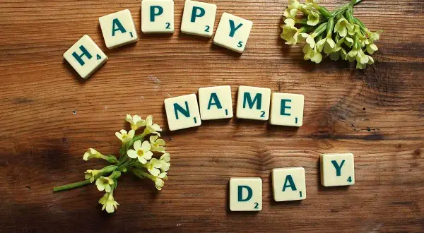 Tessere del gioco compongono "HAPPY NAME DAY" su un tavolo di legno, con fiori gialli che adornano la scena.