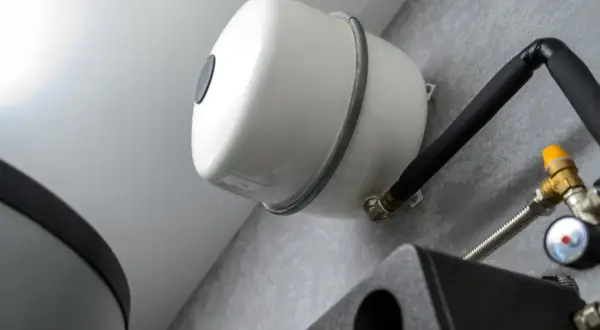 Vue rapprochée du **vase d'expansion chauffage**, élément blanc fixé au mur, avec tuyauterie noire isolée et raccords en laiton.