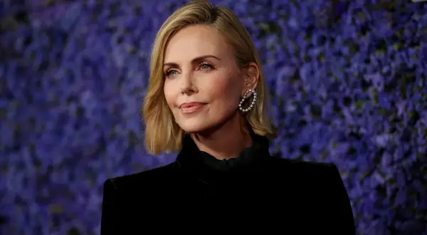 Charlize Theron, bekannt für ihre Rollen in Filmen wie "Mad Max: Fury Road", posiert vor einem lila Hintergrund.