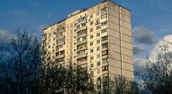 Blok mieszkalny z balkonami, otoczony drzewami. Może symbolizować służebność mieszkania, gdzie każdy balkon to osobna przestrzeń.