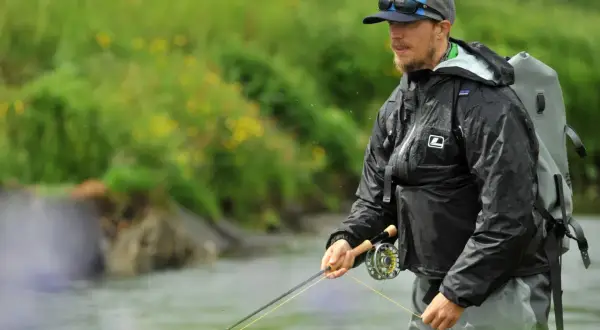 Ein Mann mit Hut und Sonnenbrille steht im Wasser und übt das **streamer fischen**. Er trägt eine schwarze Regenjacke und einen Rucksack.