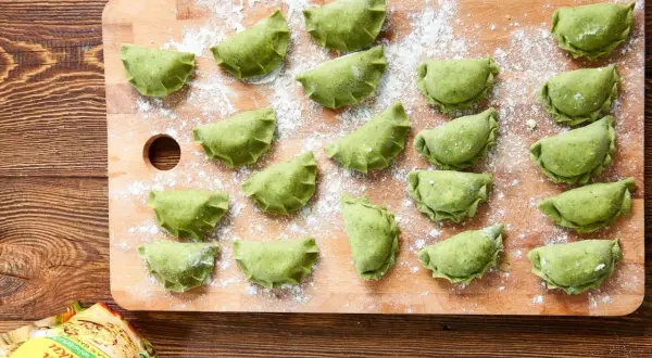 Zielone pierogi ze szpinakiem i serem feta ułożone na desce, posypane mąką.