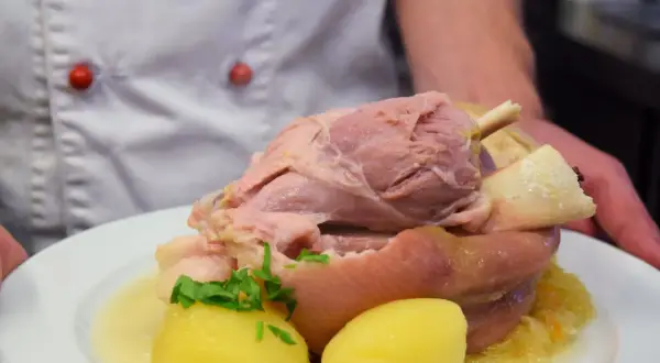 Ein Koch präsentiert ein deftiges Eisbein mit Kartoffeln und Sauerkraut. Die Frage "Eisbein wie lange kochen?" beschäftigt viele Hobbyköche.