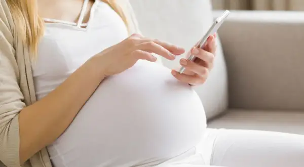 Kobieta w 24. tygodniu ciąży przegląda coś na telefonie, siedząc na kanapie.