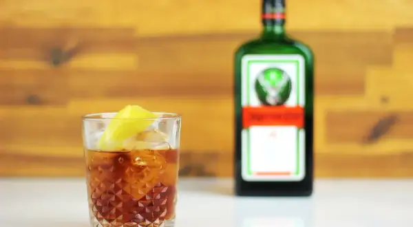 Orzeźwiający jager z colą, z plasterkiem cytryny, obok zielonej butelki Jägermeistera.