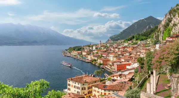 Vista panoramica di una città con tetti rossi e case colorate, incastonata tra la montagna e il blu del lago di Garda. Una nave solca le acque tranquille.