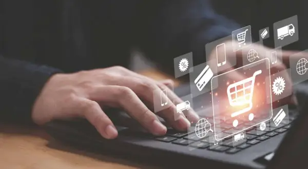 Dłoń na klawiaturze laptopa, nad nią ikony symbolizujące e-commerce: koszyk, karta płatnicza, ciężarówka, procenty, globus.