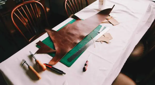 Sur une table recouverte d'une toile blanche, des outils de maroquinerie et des pièces de cuir sont disposés. On y voit un grand morceau de cuir brun, une planche à découper verte, une règle, un cutter, et des outils pour travailler le cuir, suggérant ...