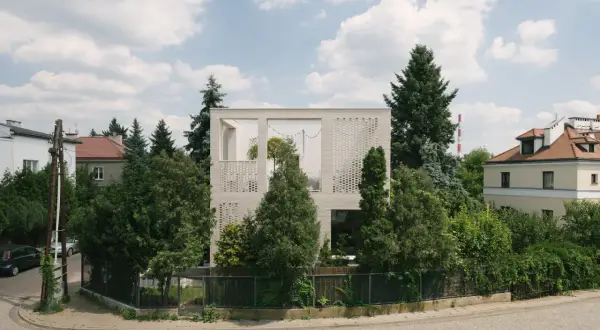 Nowoczesny dom typu kostka z białej cegły, otoczony zielenią. Widać fragmenty sąsiednich budynków i błękitne niebo z chmurami.