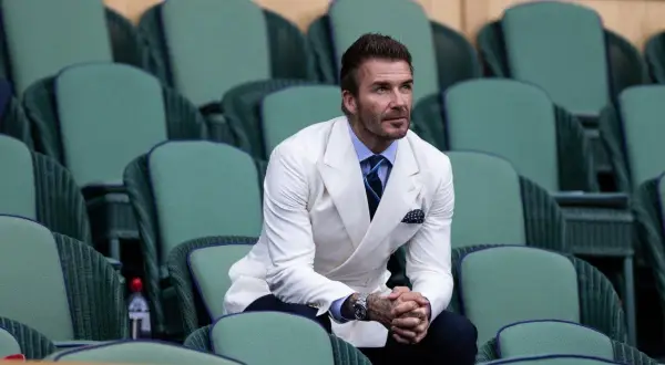 David Beckham in una giacca bianca uomo abbinamento impeccabile, con camicia azzurra e cravatta blu, siede tra le tribune verdi di Wimbledon.