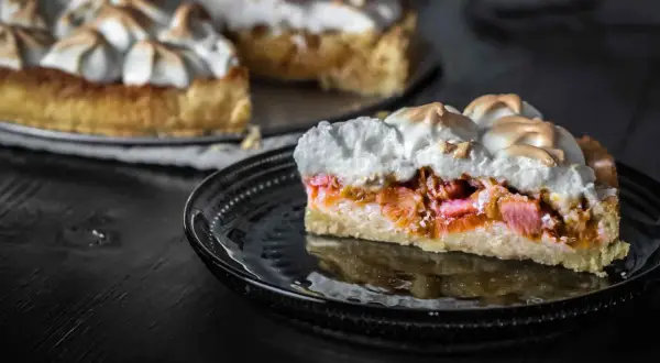 Ein Stück Rhabarberkuchen mit Baiser, das auf einem schwarzen Teller liegt. Der Kuchen hat eine goldbraune Kruste und eine saftige Rhabarberfüllung.