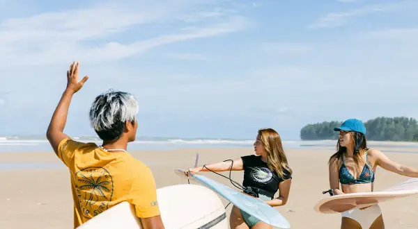 Tre vänner med surfbrädor på stranden i Thailand. En man med vitfärgat hår vinkar, medan två kvinnor förbereder sig för att surfa.