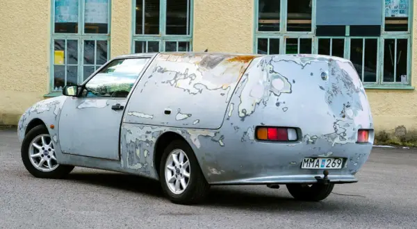En sliten Saab 600, med rostfläckar och avskavd lack, står parkerad framför en gul byggnad med gröna fönster.