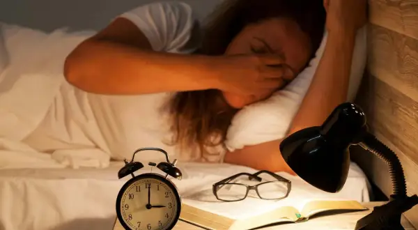 En person som gnuggar sig i ögonen i sängen, med en väckarklocka, glasögon och en bok på nattduksbordet. Känns som att vaknar flera gånger per natt.