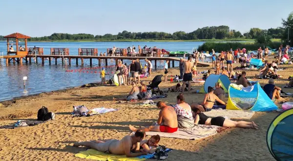 Plaża nad jeziorem powidzkim tętni życiem. Ludzie odpoczywają na piasku, kąpią się i spacerują po molo.