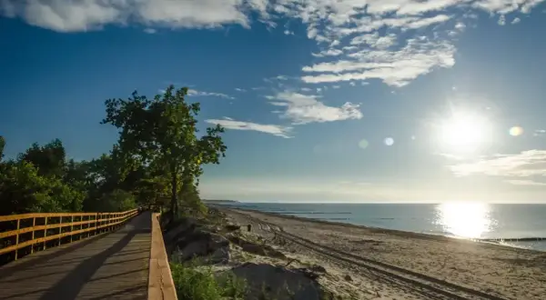 Drewniana kładka wzdłuż plaży, idealna na spacer trasą R10. Słońce odbija się w spokojnym morzu, a niebo zdobią białe chmury.