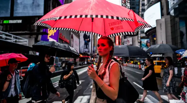 A woman with braided hair holds a bright red umbrella in this 24mm street photography shot. Rain falls on Times Square as people cross the street.