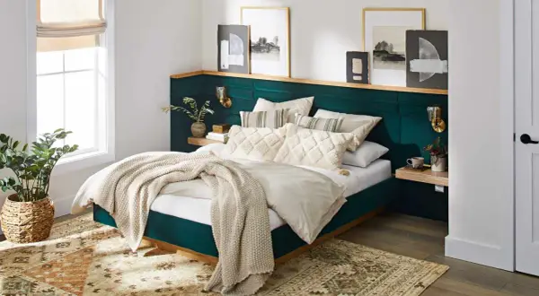 A plush, emerald green headboard with a wooden shelf above offers unique headboard ideas. Artwork and sconces adorn the wall.