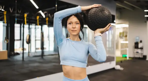 Kobieta w niebieskim stroju sportowym trzyma czarną piłkę nad głową. To ćwiczenie, które może być częścią treningu hyrox.