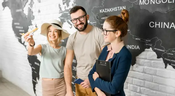 Para z paszportem i modelką samolotu przed mapą świata, obok pracownicy biura podróży.