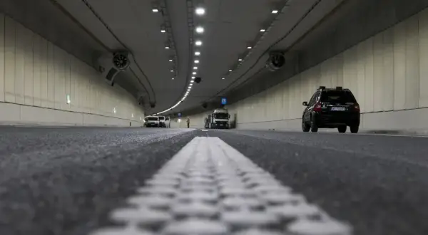 Wnętrze tunelu z widocznym ruchem pojazdów i pracami drogowymi. Widać białą linię pasów ruchu i czarny samochód. Odstęp w tunelu jest wyraźnie zaznaczony.