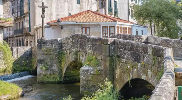 Puente de piedra con arcos sobre río, cruz de piedra y edificios históricos. Un lugar encantador que ver en Caldas de Reis.