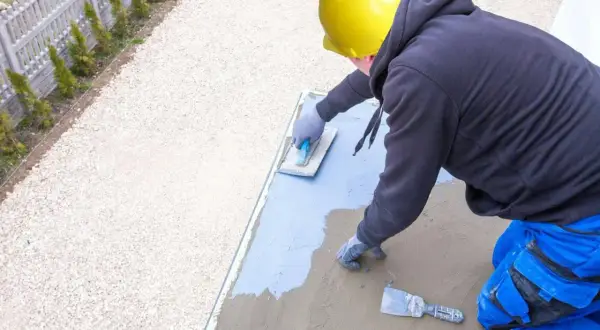 Pracownik w żółtym kasku wykonuje hydroizolację tarasu, nakładając masę izolacyjną na betonową płytę.