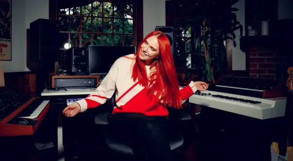 Noonie Bao, with vibrant red hair, smiles and gestures in a music studio filled with synthesizers and speakers.