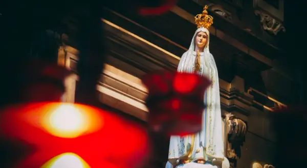 Figurka Matki Bożej Fatimskiej w koronie, z rękami złożonymi do modlitwy. W tle widać architekturę kościoła, a na pierwszym planie rozmyte czerwone światła, symbolizujące trzecia tajemnica fatimska.