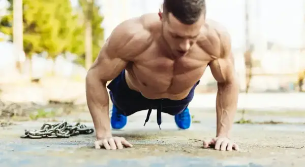 Mięśniowy mężczyzna wykonuje pompki na biceps na zewnątrz. Obok leży ciężki łańcuch.