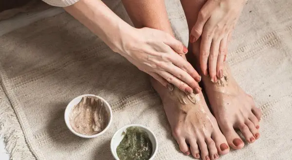 Kobieta aplikuje na stopy peeling, przygotowując je do użycia skarpetek złuszczających. Dwie miseczki z kosmetykami stoją obok.