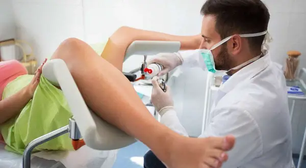 Lekarz bada pacjentkę, która leży na fotelu ginekologicznym. Widoczne są nogi pacjentki i narzędzia medyczne. Obraz pokazuje, jak wygląda zszyte krocze po porodzie.