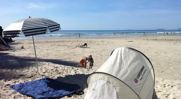 Na plaży rozłożony namiot i koc, idealne miejsce, by sprawdzić, czy można spać na plaży. Dzieci bawią się w piasku, a w tle widać morze.