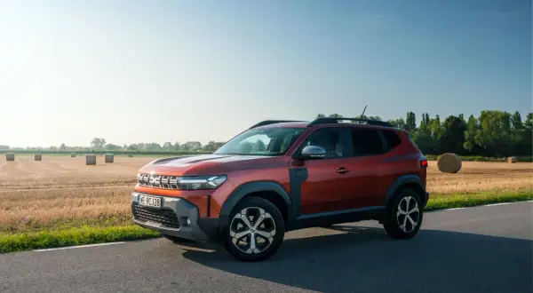 Nowa Dacia Duster w kolorze rudym, zaparkowana na poboczu drogi, z polem słomy w tle.