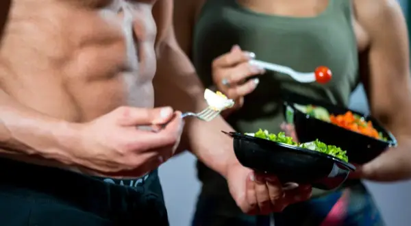 Uomo con addominali scolpiti mangia un uovo sodo, parte di una dieta ipocalorica uomo. Accanto, una donna con un'insalata.